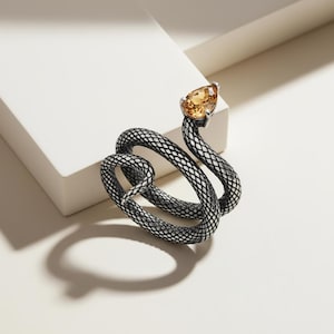 May include: A silver snake ring with a textured, scale-like pattern. The snake's head features a pear-shaped, amber gemstone. The ring is displayed on a white surface, highlighting its intricate design and colour contrast.