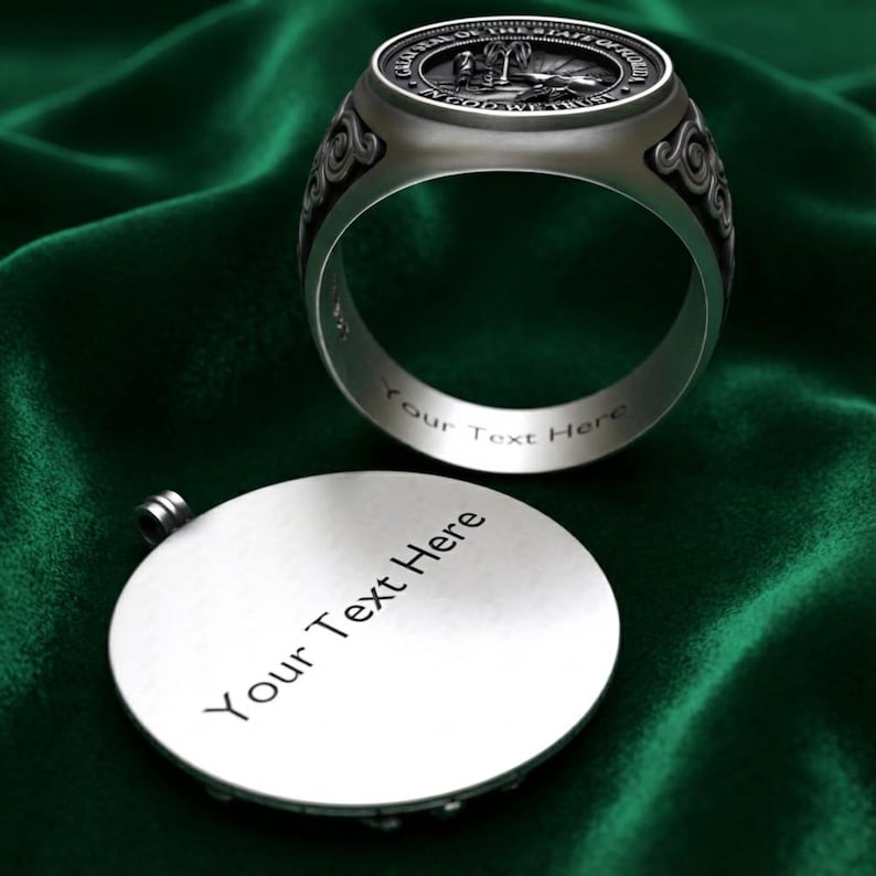 A silver ring with a decorative band and a round metal plaque with text on it, both placed on a green fabric background.