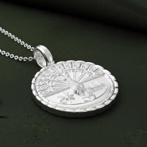 May include: A silver pendant necklace featuring a detailed peacock design. The round pendant has an intricate border and a detailed peacock in the center. The necklace is on a silver chain, and the background is a dark green.
