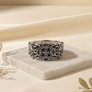 Könnte beinhalten: Ein silberner Ring mit einem verzierten Design, das ein zentrales Kreuzmotiv umgeben von wirbelnden Mustern aufweist. Der Ring wird auf einer weißen Marmoroberfläche präsentiert, die seine komplizierten Details und seine Handwerkskunst zeigt.