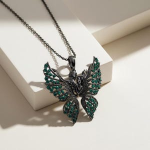May include: A black metal butterfly pendant necklace with green gemstones. The pendant features a detailed design of a figure with outstretched wings, and is suspended from a matching chain. The butterfly wings are adorned with emerald-colored stones.