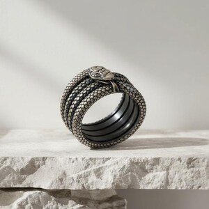 Op de afbeelding: Een zilverkleurige ring in de vorm van een opgerolde slang. De ring heeft een gedetailleerde slangenkop en schubben, met meerdere banden die het lichaam van de slang vormen. De ring wordt gepresenteerd op een getextureerd, licht oppervlak.