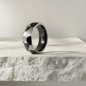 May include: A geometric design ring with a faceted exterior. The ring features a silver-colored exterior with black accents and a black interior. The ring is displayed on a white, textured surface.