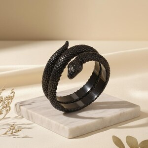 May include: A black snake-shaped ring, coiled around itself, resting on a marble block. The ring has a textured surface, with the snake's head and scales clearly defined. The background is a soft, neutral color.