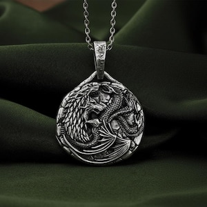 Op de afbeelding: Een zilveren hangerketting met een gedetailleerd drakenontwerp. De hanger heeft een cirkelvorm met twee draken die tegenover elkaar staan. De ketting heeft een zilveren ketting, tegen een donkergroene stoffen achtergrond.
