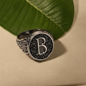Könnte beinhalten: Ein silberner Siegelring mit einem verzierten, wirbelnden Muster um das Band. Die Vorderseite des Rings zeigt den Buchstaben "B" in einer fetten Großschrift, vor einem dunklen Hintergrund. Der Ring wird auf einer neutralfarbenen Oberfläche präsentiert.
