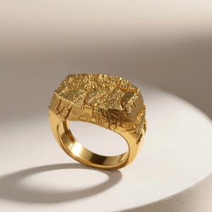 May include: A gold-colored ring with a textured, rectangular top. The ring has a thick band and a detailed, rough surface on the top, resembling a rock formation. The ring is displayed on a white surface.