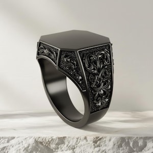 Puede incluir: Un anillo de sello negro con una cara octogonal y detalles florales ornamentados. El anillo está hecho de un material oscuro y mate y descansa sobre una superficie blanca texturizada. El diseño del anillo presenta intrincados patrones arremolinados.