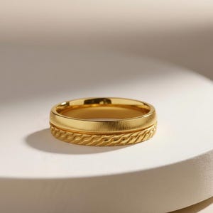 May include: A gold ring with a textured band and a smooth upper band. The ring is set against a white background with soft lighting, highlighting the ring's metallic sheen. The ring is a piece of jewelry.