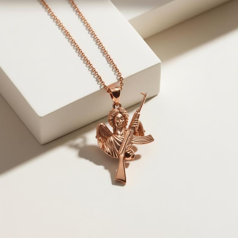 Puede incluir: Collar de color oro rosa con un colgante que representa un &aacute;ngel sosteniendo un rifle. El &aacute;ngel tiene alas detalladas y una t&uacute;nica fluida. La cadena es delicada. El collar se exhibe sobre una superficie blanca.
