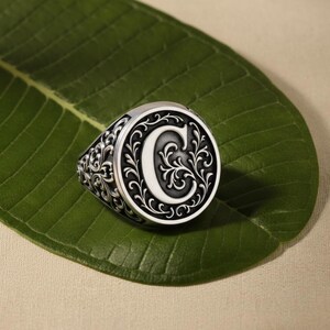 Könnte beinhalten: Ein silberner Ring mit einer großen, kreisförmigen Oberfläche, die den Buchstaben "C" in Weiß zeigt, umgeben von einem kunstvollen, wirbelnden schwarzen Design. Der Ring hat ein detailliertes Blumenmuster auf dem Band. Der Ring liegt auf einem grünen Blatt.