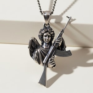 Puede incluir: Collar con colgante plateado que representa a un &aacute;ngel sosteniendo un rifle de asalto. El &aacute;ngel tiene alas detalladas y cabello suelto. La cadena del collar es un dise&ntilde;o de cuerda trenzada. El colgante es un dise&ntilde;o detallado en tres dimensiones.