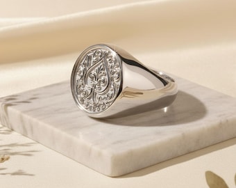 Puede incluir: Un anillo de sello plateado pulido con una cara ovalada que muestra un elaborado motivo de pica. El anillo está colocado sobre una superficie de mármol. El diseño es detallado e intrincado, lo que refleja un estilo clásico.