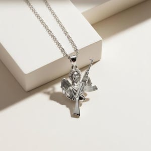 Puede incluir: Un collar con colgante plateado que representa un &aacute;ngel sosteniendo un rifle. El &aacute;ngel tiene alas y rasgos faciales detallados. La cadena del collar tambi&eacute;n es plateada y tiene un dise&ntilde;o de eslabones. El colgante es un dise&ntilde;o tridimensional detallado.