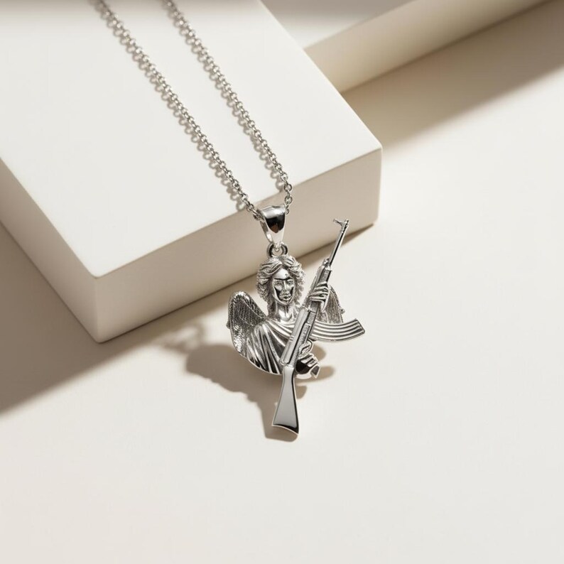 Puede incluir: Un collar con colgante plateado que representa un &aacute;ngel sosteniendo un rifle. El &aacute;ngel tiene alas y rasgos faciales detallados. La cadena del collar tambi&eacute;n es plateada y tiene un dise&ntilde;o de eslabones. El colgante es un dise&ntilde;o tridimensional detallado.