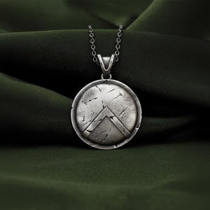 Puede incluir: Un collar con colgante plateado con un diseño de escudo redondo y un galón. El escudo tiene un aspecto desgastado y texturizado con grietas. El collar cuelga de una delicada cadena, sobre un fondo verde oscuro.