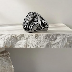 May include: A silver-toned ring featuring a detailed depiction of an Egyptian pharaoh's face. The ring has an oval face with intricate patterns and symbols on the band. The pharaoh's headdress is adorned with small, dark accents.