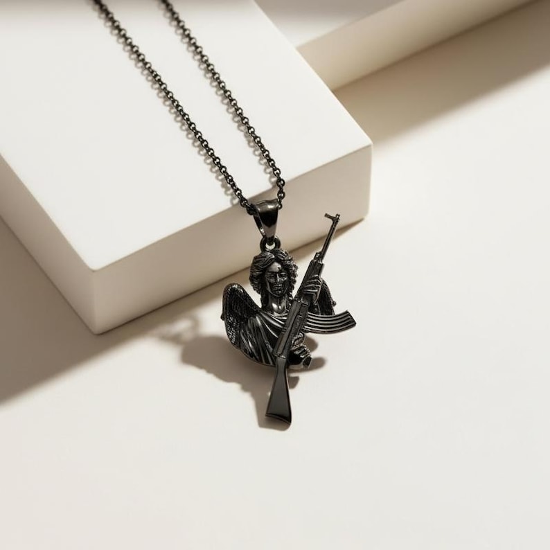 Puede incluir: Collar de metal negro con un colgante de &aacute;ngel sosteniendo un rifle. El colgante tiene alas y rasgos faciales detallados. La cadena es negra, creando un accesorio audaz y llamativo.
