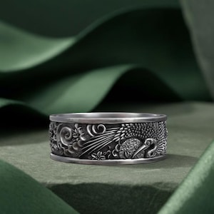 Puede incluir: Un anillo plateado con un diseño grabado que presenta una grulla, nubes y elementos florales. El anillo tiene una superficie detallada y texturizada y una anchura de banda de aproximadamente 0,8 cm. El anillo se muestra sobre una superficie verde.