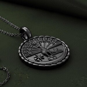 May include: A black pendant necklace featuring an oval-shaped pendant with a detailed design. The pendant showcases a bird with a fanned tail, a building, and a starburst design. The necklace chain is also black.