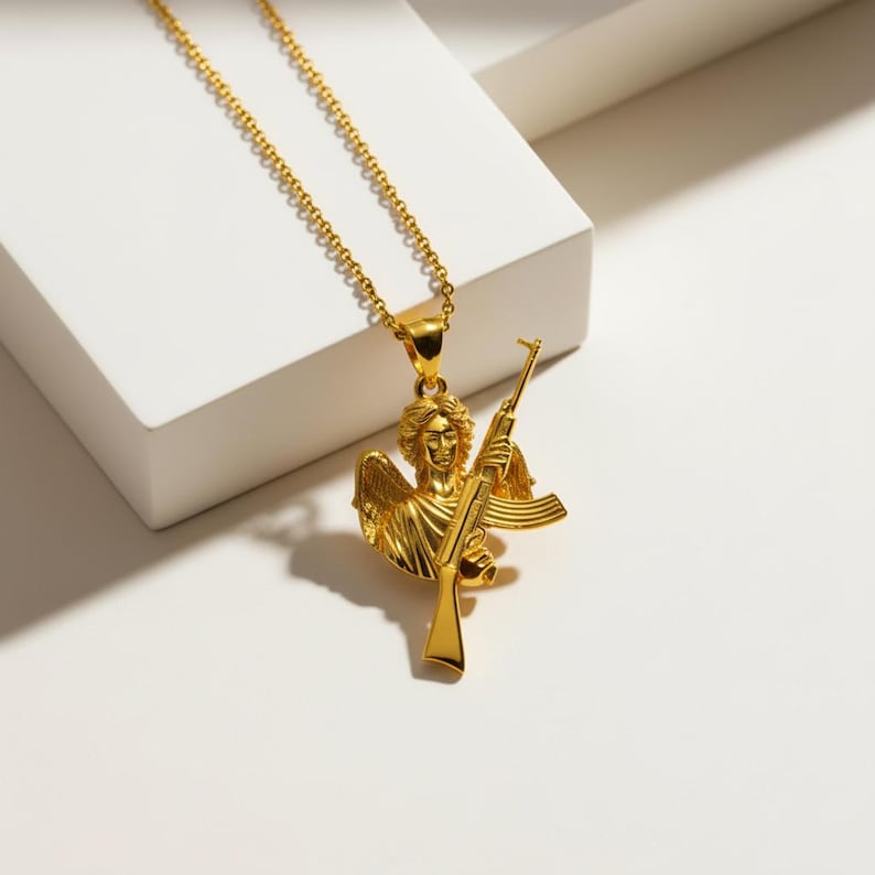 Puede incluir: Collar con colgante dorado que representa un &aacute;ngel sosteniendo un rifle. El &aacute;ngel tiene alas y rasgos faciales detallados. La cadena del collar tambi&eacute;n es dorada y cuelga sobre una superficie blanca.