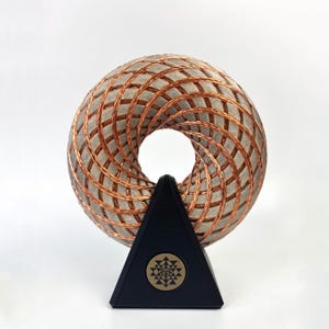 Peut inclure: Sculpture géométrique en forme de tore, composée de blocs gris et de fil de cuivre. Elle repose sur un socle triangulaire noir avec un emblème circulaire. Design moderne et abstrait, pour la décoration.