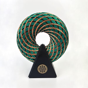 Può includere: Una scultura a forma di toroide verde e color rame. Il design intrecciato presenta un motivo a rombi ripetuto. È montata su una base triangolare nera con un emblema color oro. La scultura è esposta su uno sfondo bianco.