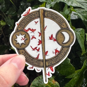 May include: A circular sticker featuring a bronze-colored frame with a vertical pin, a sun icon on the left, and a crescent moon on the right. The text PEREGRINI HURURAE is inscribed on the frame. The center contains scattered red bird silhouettes.