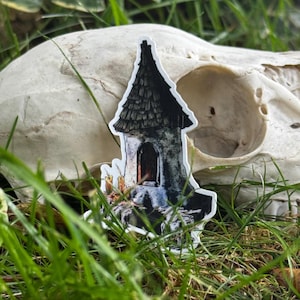 May include: A die-cut sticker featuring a tall, narrow stone tower with a dark, textured shingled roof. The tower has a single arched doorway with a warm orange glow inside and a cluster of small, lit candles placed at the base of the structure.