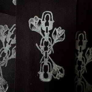 May include: Close-up of black fabric patches with a repeating silver floral and lock design. The patches are rectangular and feature a detailed, stylized pattern. The design includes floral elements and a series of connected locks.