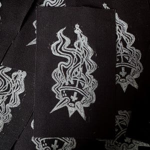 May include: Black rectangular fabric patches with a silver-colored graphic of a crown engulfed in flames. The patches are densely arranged, creating a repeating pattern. The design features a star and wavy lines.