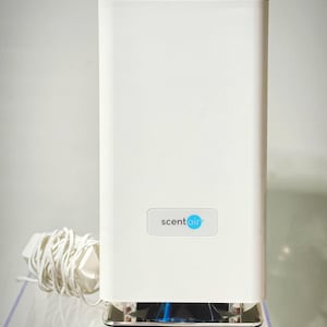 May include: A white rectangular scent diffuser with the text "scentair" on the front. The diffuser has a silver base and a white power cord. The top of the diffuser has a series of small openings.