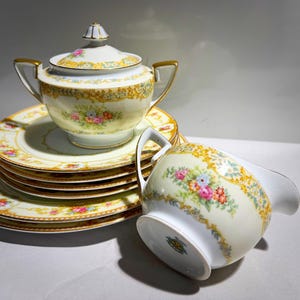 Puede incluir: Un juego de té de porcelana vintage con azucarero con tapa, jarra para crema y varios platos. El juego tiene una base color crema con detalles florales y dorados. Los platos están apilados y la jarra de crema está inclinada.