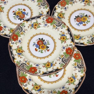 May include: A set of four vintage ceramic plates. Each plate features a cream-colored center with a floral bouquet and a decorative border of orange, yellow, and blue flowers. The rims have a black and brown patterned design.