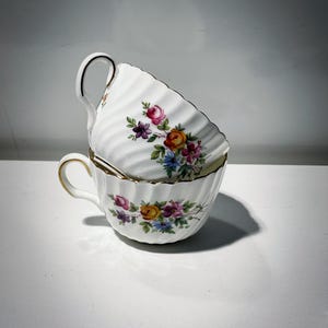 May include: Two white teacups stacked, each adorned with a floral pattern in pink, yellow, purple, and blue. The cups have a ribbed texture and gold trim on the rim and handle, creating an elegant aesthetic.