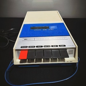 以下が含まれることがあります： クリーム色の上面と青と銀色の前面パネルを備えた、ヴィンテージのカセットレコーダー。レコーダーには、いくつかの黒いボタン、赤い録音ボタン、青いキャリングストラップが付いています。「CASSETTE RECORDER」の文字が見えます。