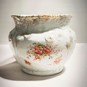 Pode incluir: Um vaso de cerâmica branco com borda ondulada e design floral. O vaso apresenta acabamentos e detalhes dourados, com um grupo de flores rosa e outros detalhes florais coloridos. O vaso tem formato bulboso.