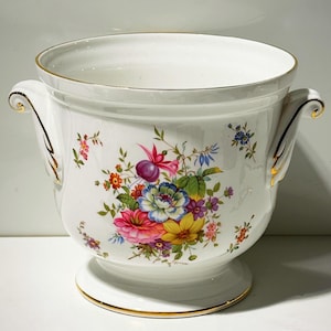 May include: A white ceramic planter with a floral design featuring pink, blue, yellow, and purple flowers. The planter has gold trim and handles. The base is round and the interior is white.
