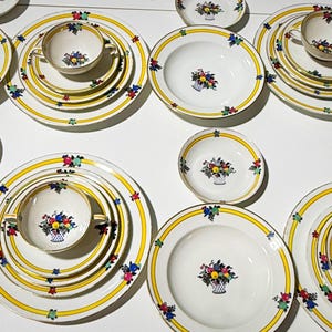 May include: A collection of white porcelain dinnerware with a yellow border and floral basket design. The set includes plates, bowls, and teacups, arranged on a white surface. The floral design features red, blue, and yellow flowers.