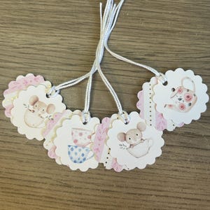May include: A collection of six scalloped-edge gift tags with white string. Each tag features a watercolor illustration of a mouse, teacup, or teapot with pink and gold accents. The tags are arranged on a wooden surface.