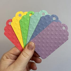 May include: A hand holds a fanned array of colorful gift tags. The tags are red, yellow, green, light blue, dark blue, and lavender. Each tag has a scalloped edge, a small hole, and a textured polka dot pattern.
