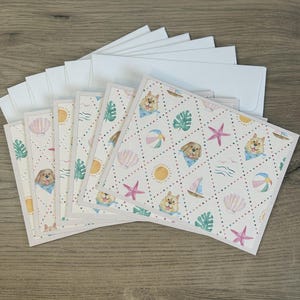 May include: A set of greeting cards and envelopes. The cards feature a repeating pattern of a dog wearing a blue bandana, seashells, starfish, and a sun on a white background. The envelopes are white.