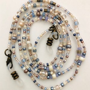May include: A beaded eyeglass chain in shades of white, blue, and pearl. The chain features a variety of small beads, including round, square, and oblong shapes. The chain has bronze-colored clasps and loops for attaching to glasses.