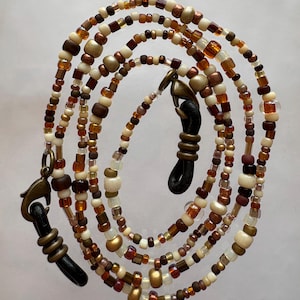 May include: Eyeglass chain with a variety of beads in shades of brown, cream, and gold. The chain has a lobster claw clasp and rubber loops to attach to glasses. The beads are various shapes and sizes.