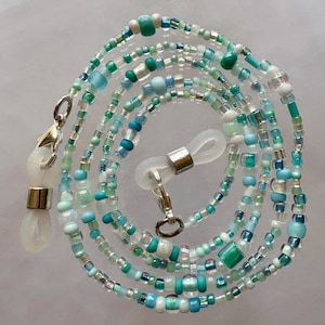May include: A beaded eyeglass chain in shades of teal, turquoise, and white. The chain is made of small, round beads and larger, square beads. Silver-colored clasps and eyeglass holders are attached to the ends.