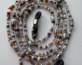 May include: An eyeglass chain featuring a variety of small glass beads in shades of white, clear, brown, gray, and metallic copper. The chain includes silver-toned metal lobster clasps attached to black rubber loops designed to secure eyewear frames.