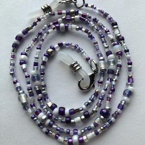 May include: A beaded eyeglass chain in shades of purple, white, and silver. The chain is made of small, round beads and larger, cylindrical beads. The chain has silver-colored clasps and clear rubber loops for attaching to glasses.