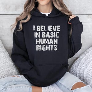 May include: Black hooded sweatshirt with the white text "I BELIEVE IN BASIC HUMAN RIGHTS". The text is in a bold, distressed font. The sweatshirt has a drawstring hood and a kangaroo pocket.