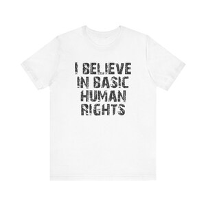 May include: White t-shirt featuring the statement "I BELIEVE IN BASIC HUMAN RIGHTS" in a distressed black font. The shirt has a crew neck and short sleeves.