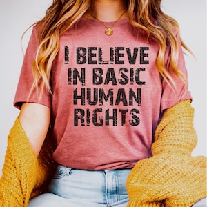 May include: A dusty rose t-shirt with the text "I BELIEVE IN BASIC HUMAN RIGHTS" in a distressed black font. The shirt is worn with a gold necklace and a yellow cardigan.